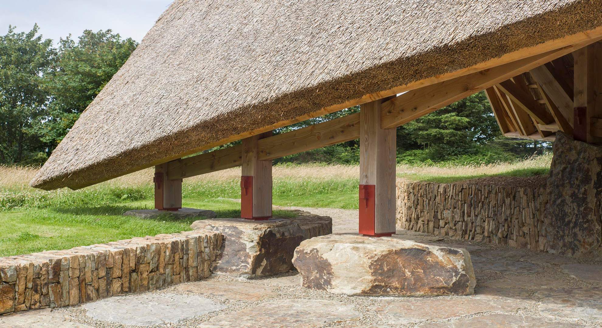 Detalle de una construcción con techo de paja, columnas de madera y base de piedra en un entorno natural.
